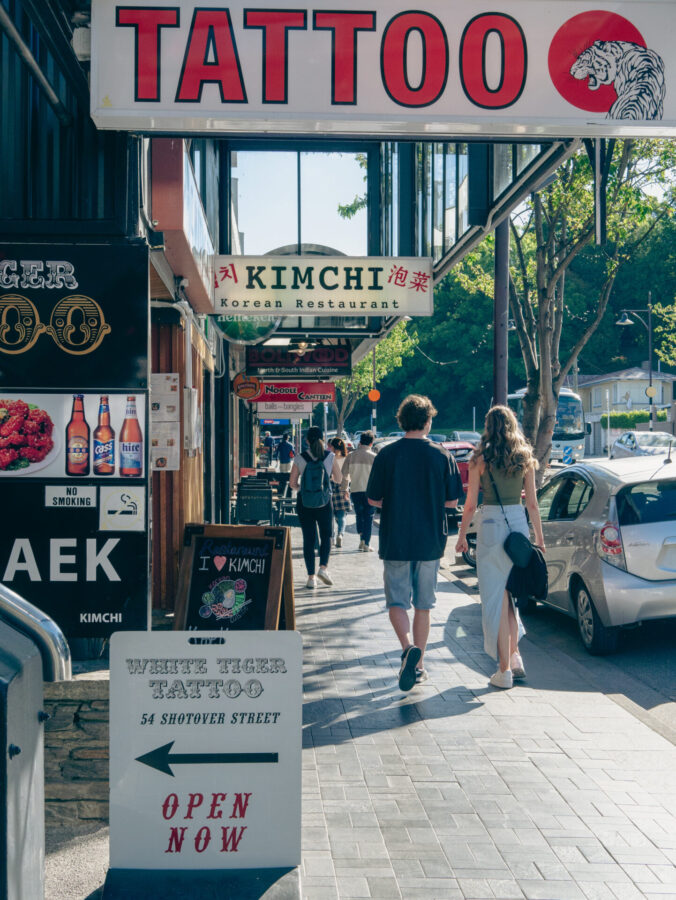 A sidewalk from a summer's day in Queenstown, New Zealand, filled with local business signs and a few pedestrians.
