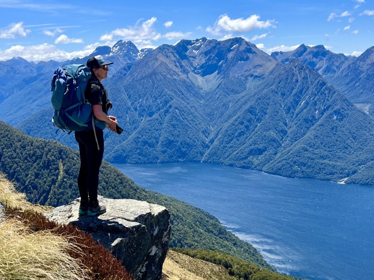A hiker standing in front of a lake and mountain scene from up along the Kepler Track's ridgeline.