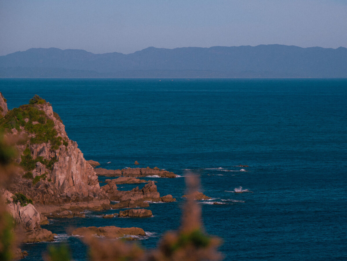 A view from up in the hills on the Abel Tasman Track in New Zealand, with the ocean, some rocky cliffs, and distant mountains in the background.